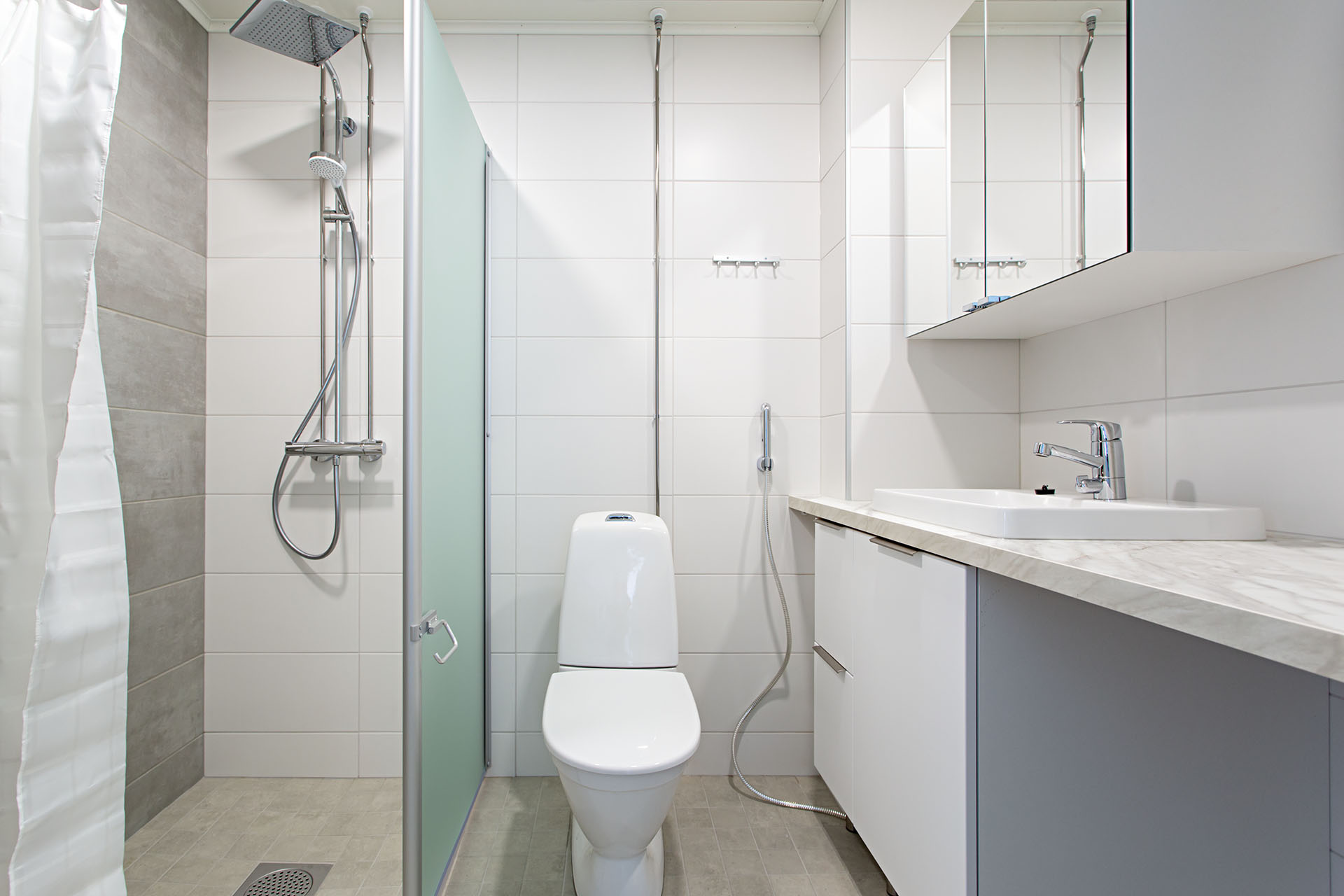 Kylpyhuone jossa keskellä WC, vasemmalla suihkutila ja oikealla lavuaari sekä peilikaappi